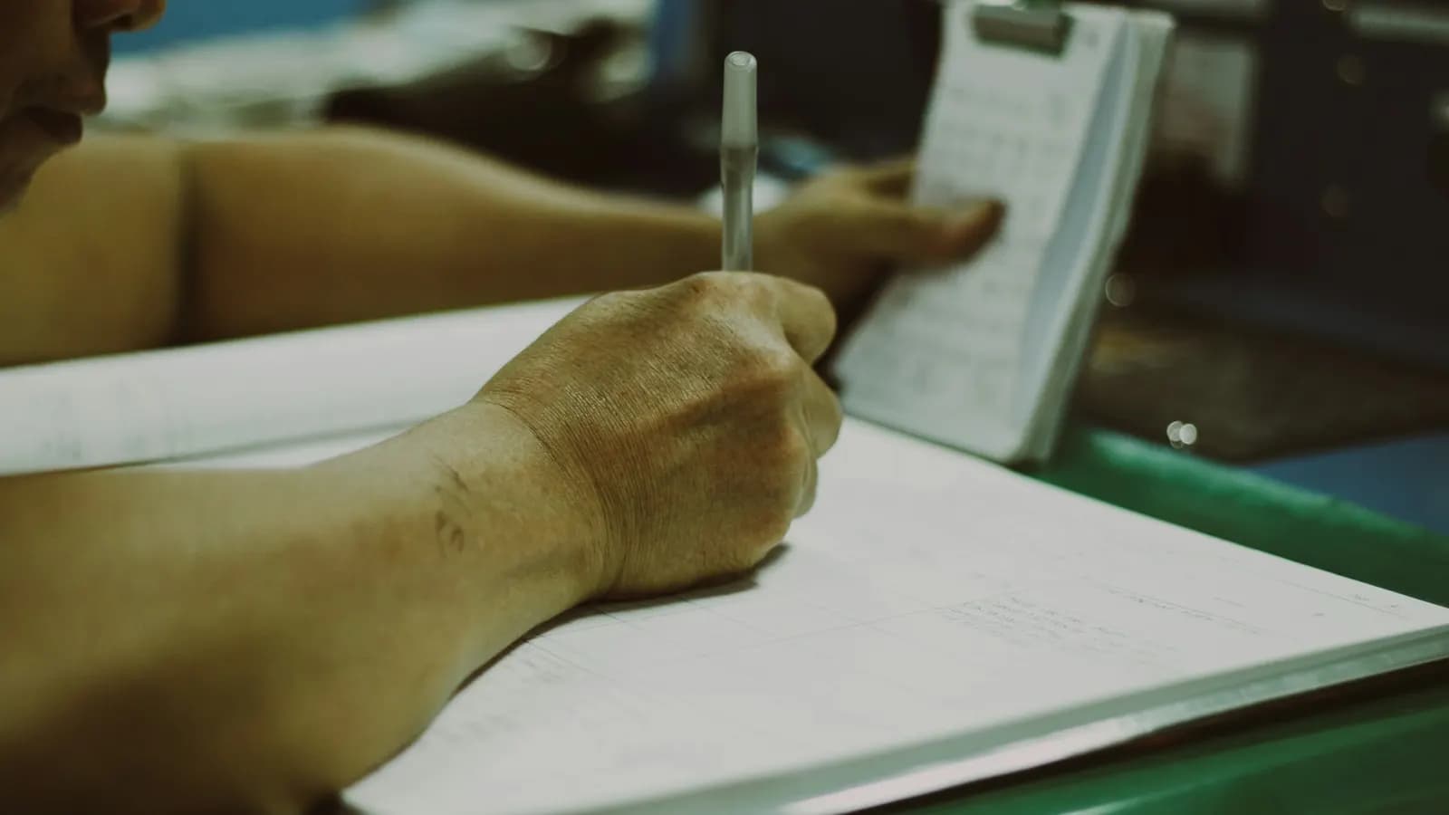 Mano che compila a penna un registro cartaceo, accanto a un blocco appunti
